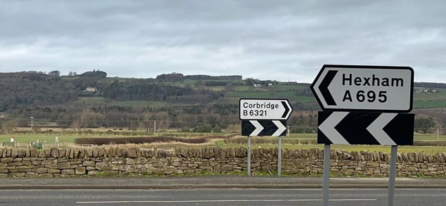 A photo at the side of the road with hills in the distance and two signs. One pointing right saying Corbridge and the other pointing left saying Hexham