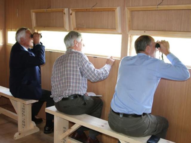 Three men in a bird watch hut sitting on a bench and looking out the viewer for bird spotting.