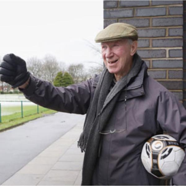 Jackie Charlton holding a football wearing a large coat and a flat cap