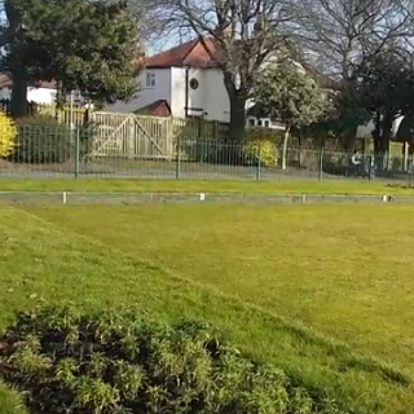 A large well maintained grass area used for bowls, with large trees in the background and a house.
