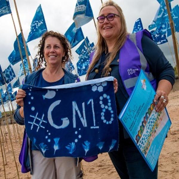 Two people on a beach holding a sun bleached flag that has #GN18 on it.