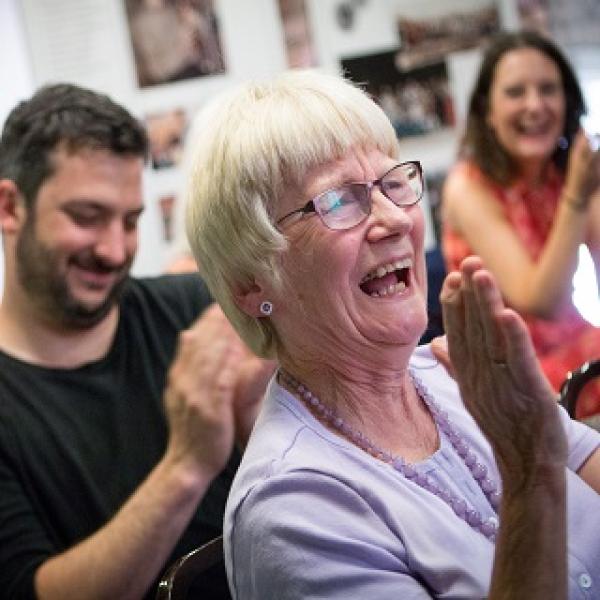 A woman with greyish hair and glasses. Laughing and smiling along while clapping her hands.