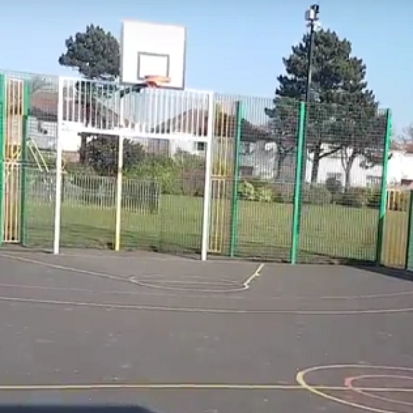 A photograph of a fenced in area with a tarmac floor painted with lines for a basketball court and a hoop at one end.
